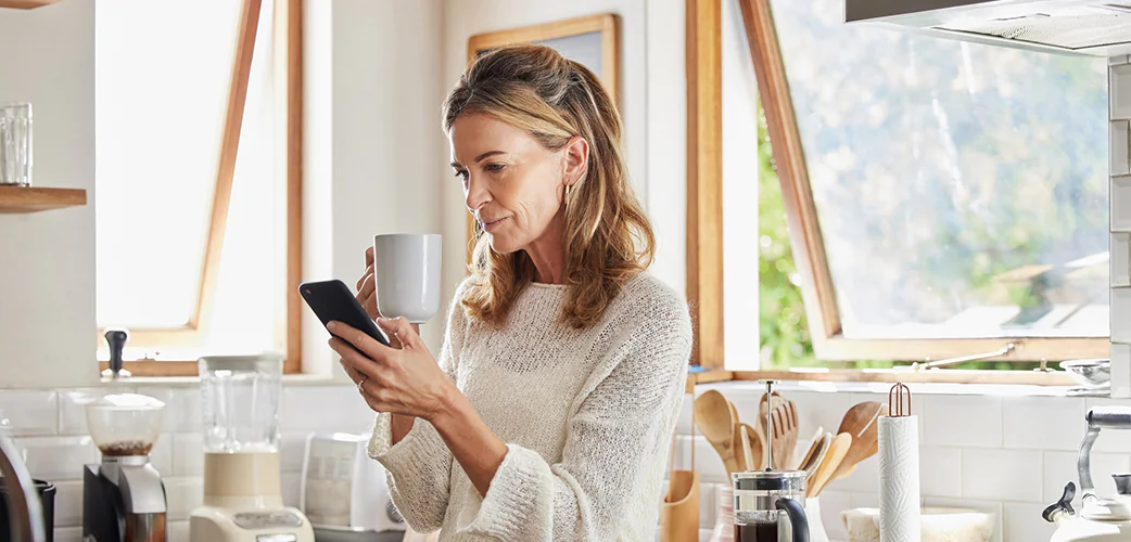 Woman in a kitchen checking her phone while holding a mug